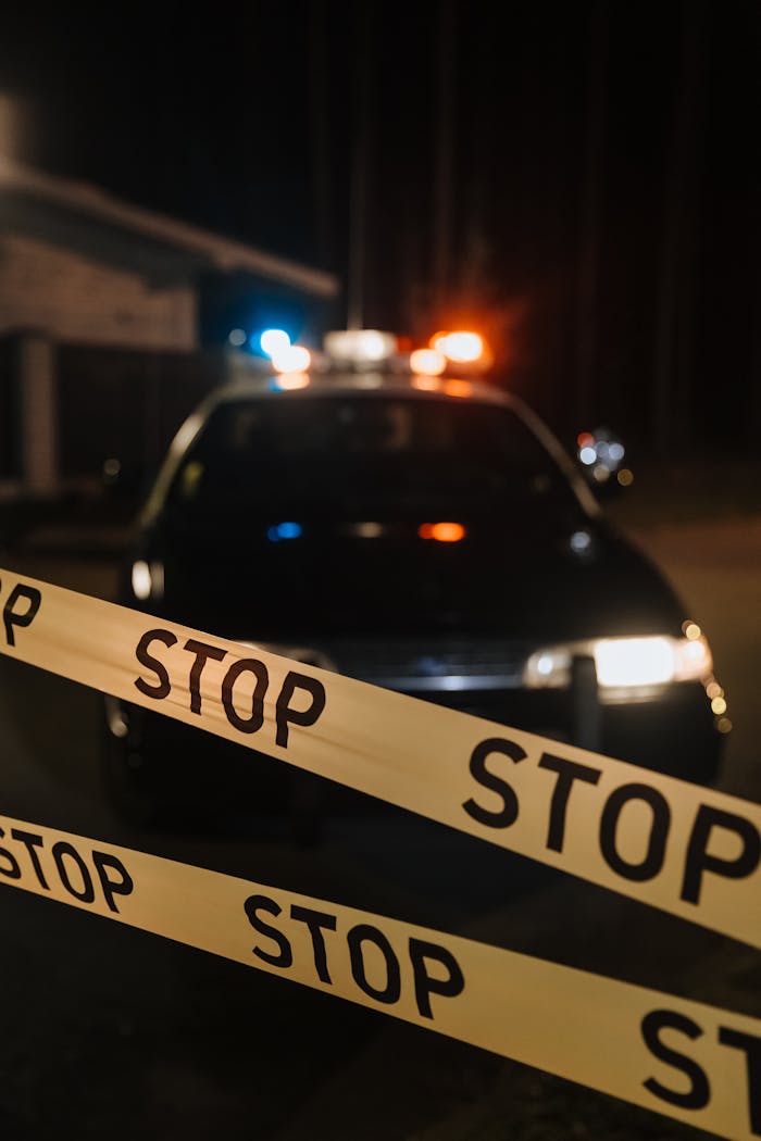 A police car with flashing lights behind a stop tape at night, depicting a crime scene.