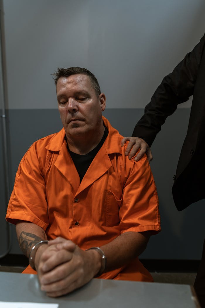 A prisoner in an orange jumpsuit sits handcuffed under supervision indoors.