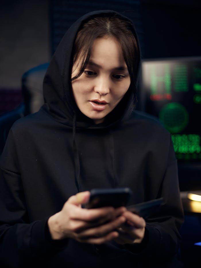 gallery-1 A young woman in a black hoodie uses her smartphone indoors, surrounded by tech gear.